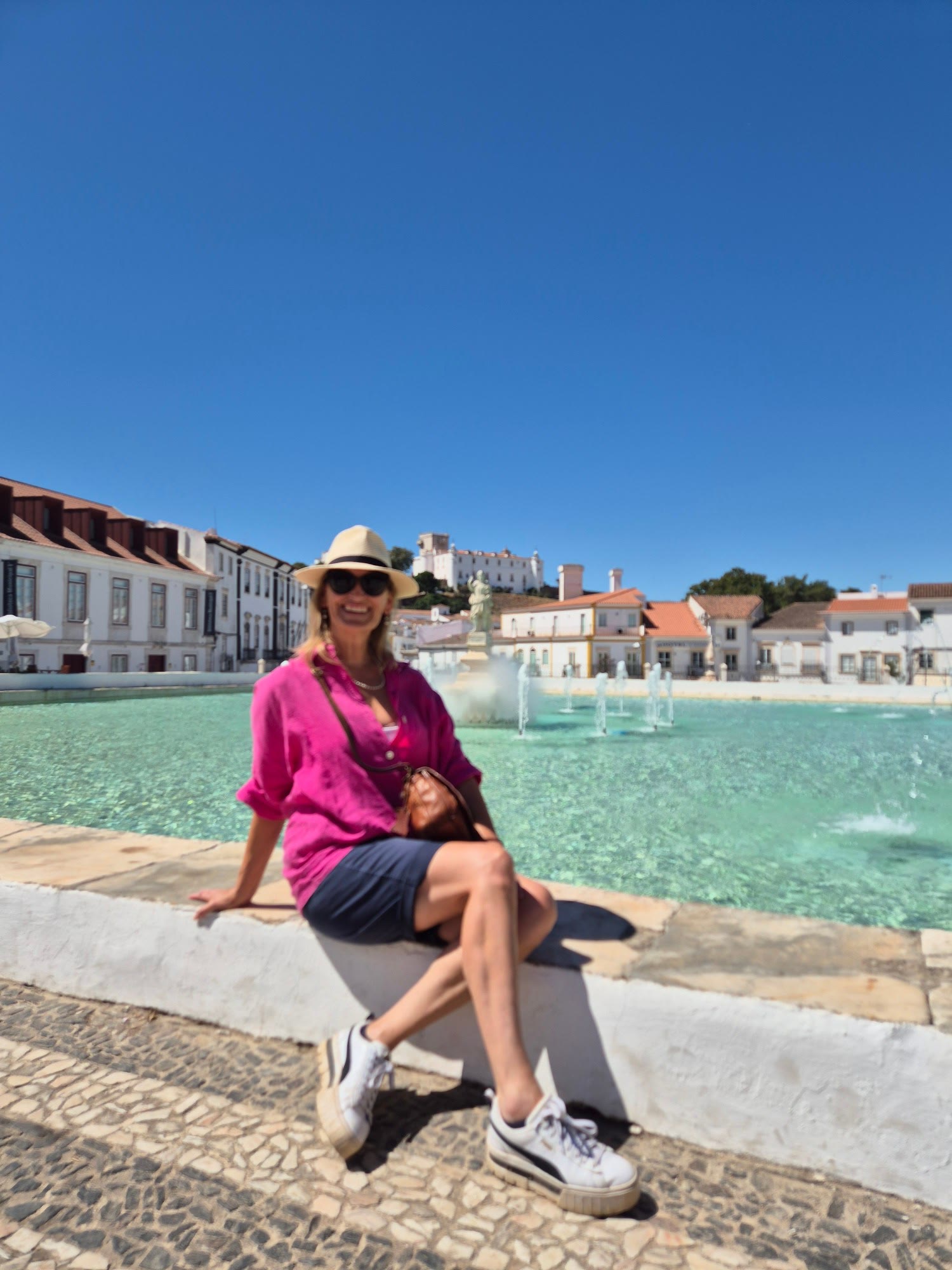 Sara in a pink shirt and straw hat, sitting by a fountain in a sunny Portuguese town square